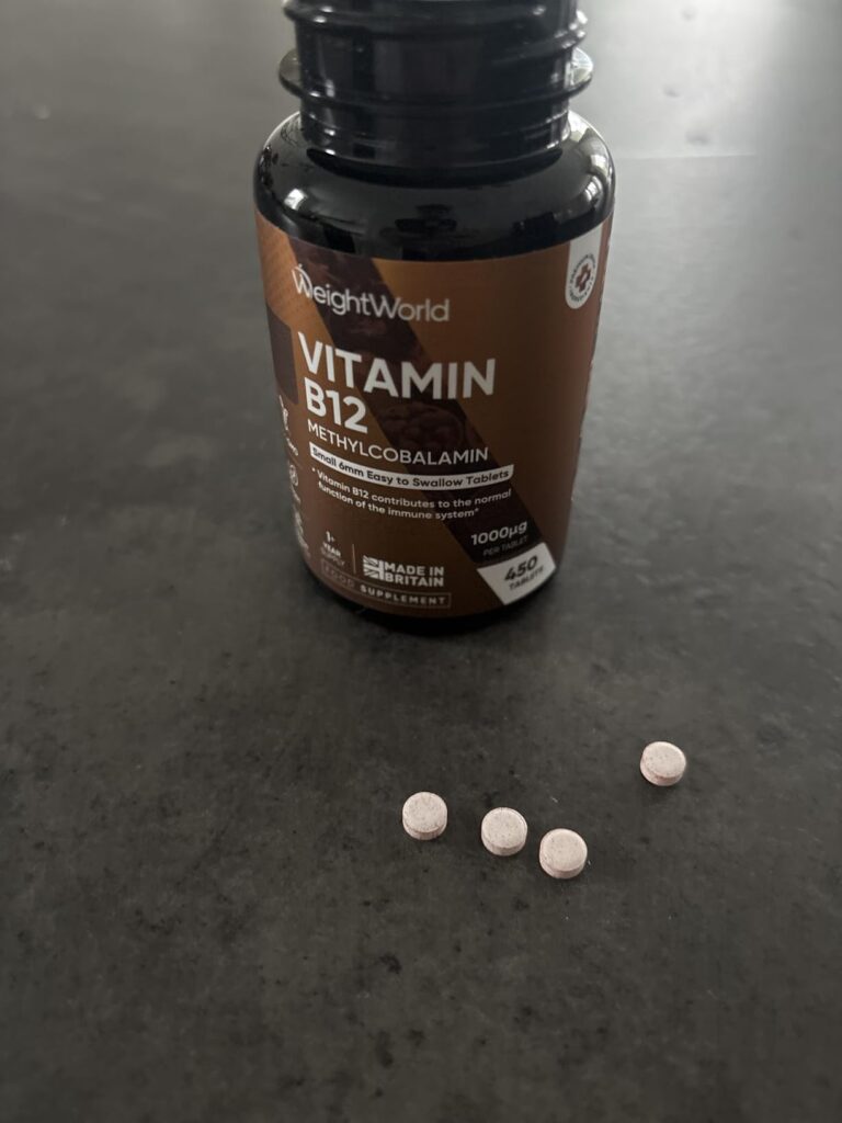 Bouteille de vitamine B12 WeightWorld et quatre comprimés roses posés sur une surface sombre.