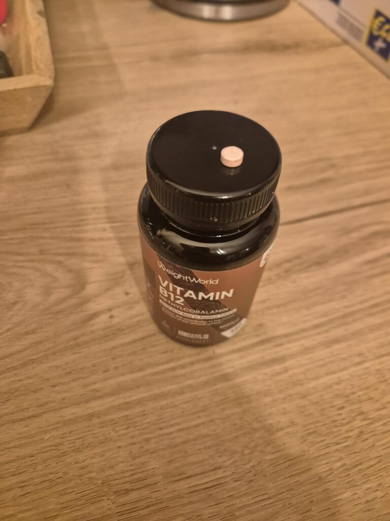 Bouteille de vitamine B12 WeightWorld posée sur une table en bois clair, avec un comprimé rose posé sur le couvercle. Angle d...
