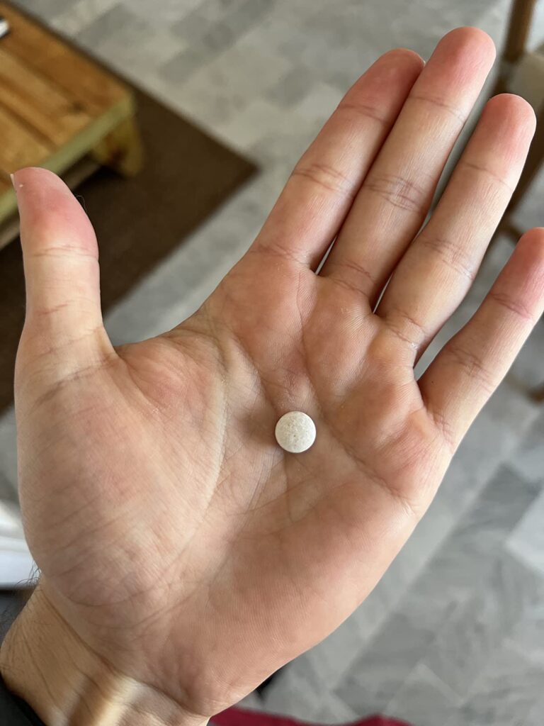 Un comprimé de multivitamines dans une main ouverte. On voit l'intérieur d'une maison en arrière-plan.