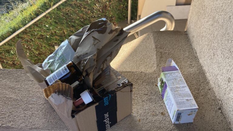 Colis Amazon avec bouteilles endommagées, posé sur des marches extérieures. Une autre boîte de produit est à côté.
