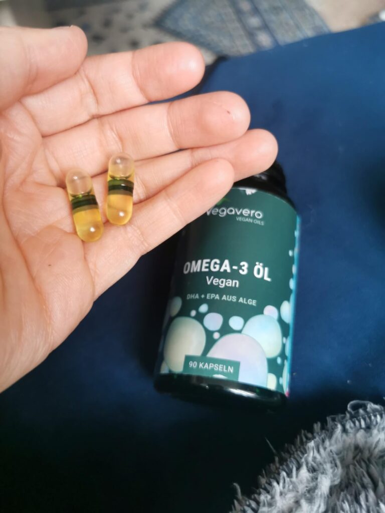 Deux gélules d'oméga-3 végétaliennes tenues dans une main devant la bouteille sur un tissu bleu, les capsules sont translucid...