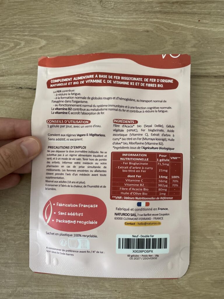 Vue de l'arrière du sachet de compléments alimentaires posé sur un plan de travail en bois clair, avec une main visible sur l...