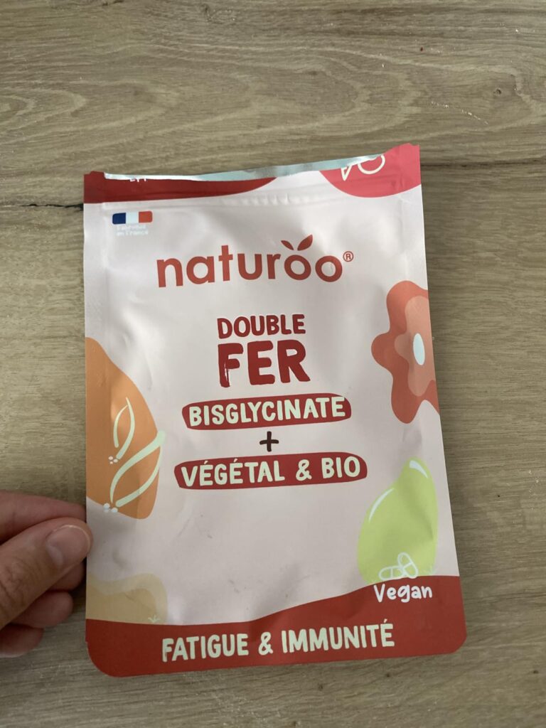 Vue de l'avant du sachet de compléments alimentaires posé sur un plan de travail en bois clair, avec une main visible sur le...