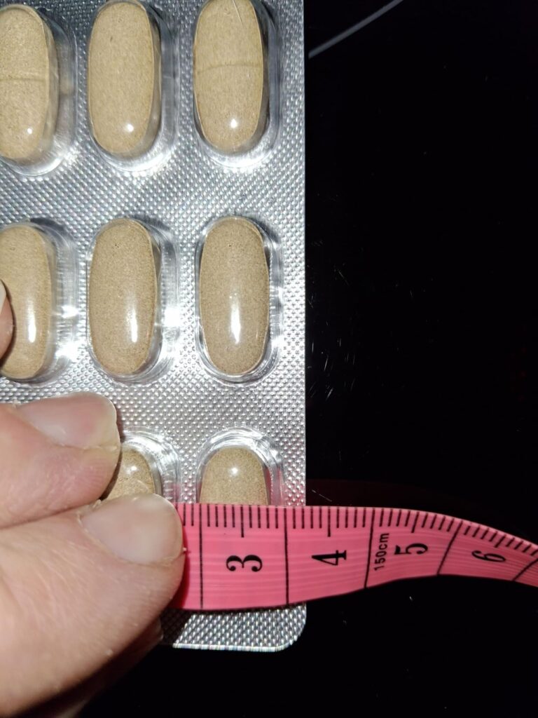 Une main tenant une capsule et un mètre ruban rose, indiquant la longueur de la capsule.