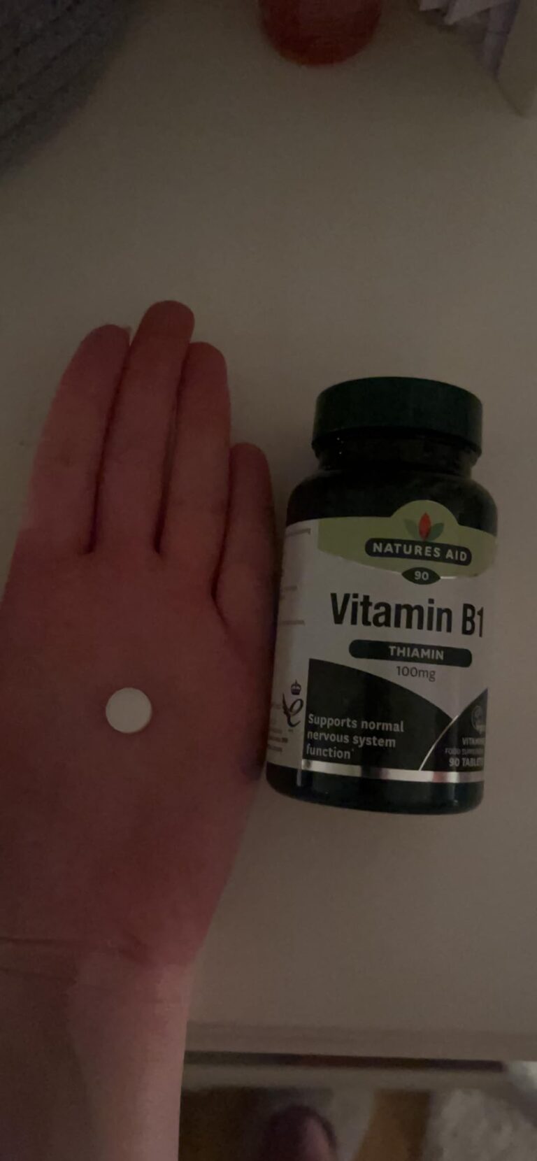 Un comprimé blanc dans une main, à côté du flacon de vitamine B1 sur une table de nuit. L'image est mal éclairée.
