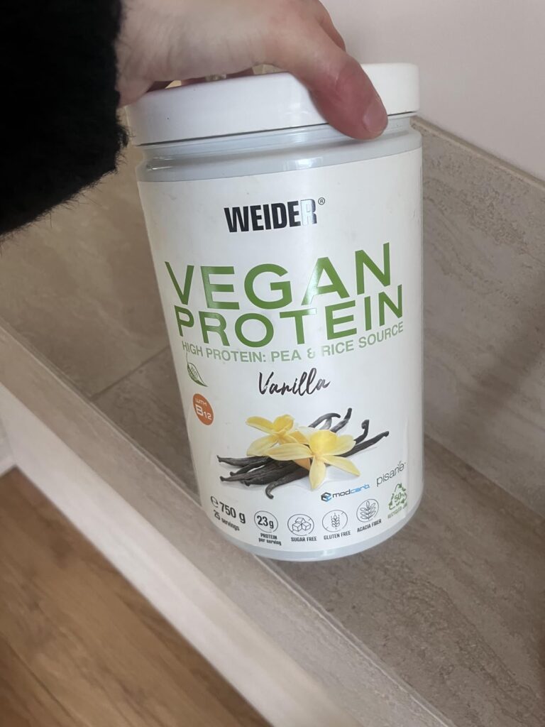 Un pot de protéine Vegan Weider saveur Vanille, tenu par une main.