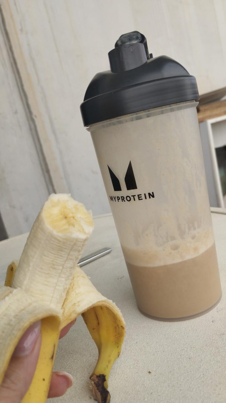 Un shaker Myprotein rempli d'une boisson protéinée, à côté d'une banane à moitié pelée. Le shaker est posé sur une surface cl...