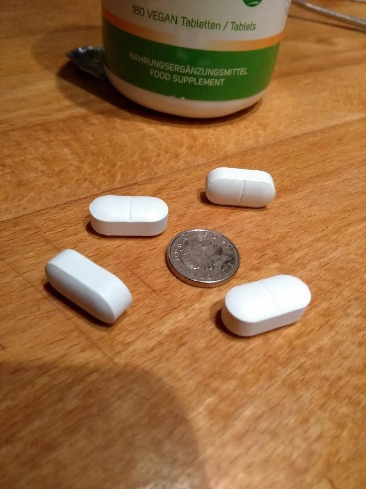 Quatre pilules blanches allongées et une pièce de monnaie pour donner l'échelle. Au fond, un flacon vert et blanc. La photo e...