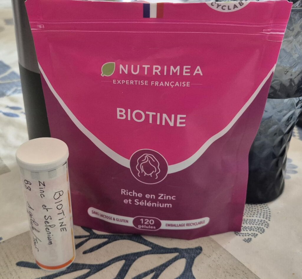 Sachet de Biotine devant un contenant cylindrique, un pot blanc et un autre sachet noir en arrière-plan
