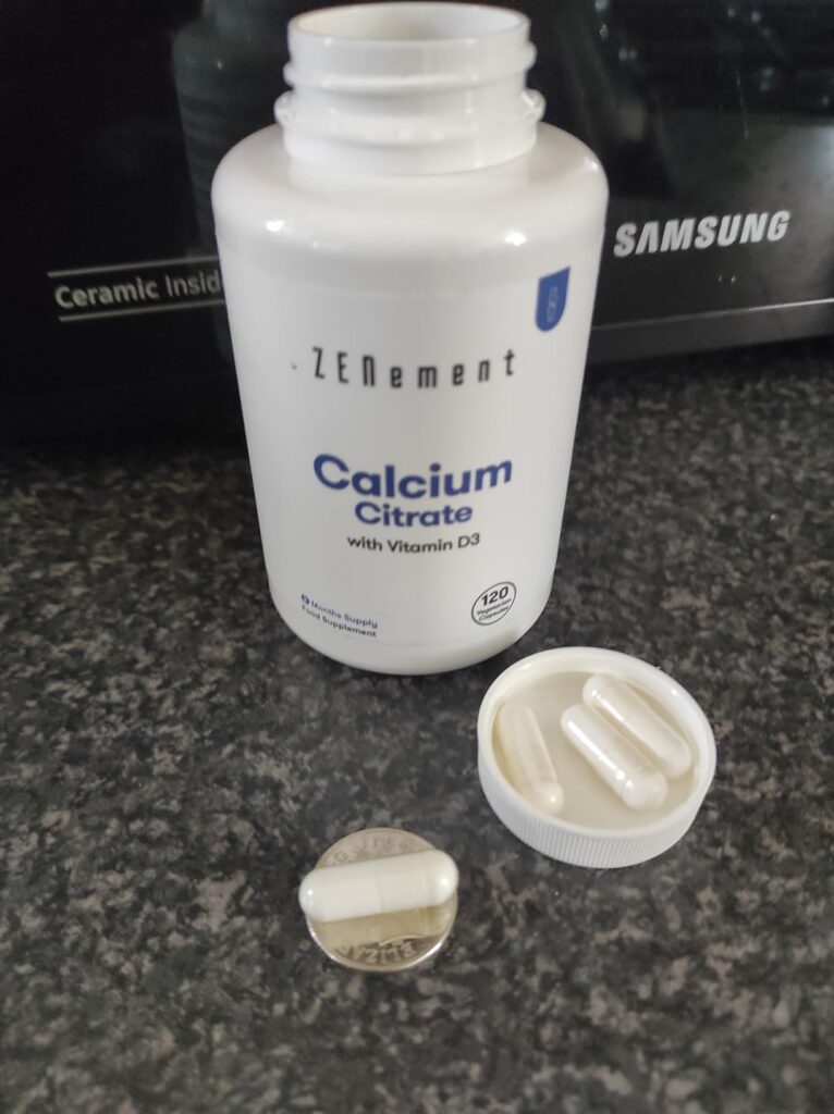 Bouteille de Calcium Citrate blanche ouverte, avec des gélules visibles dans le bouchon et sur le côté.