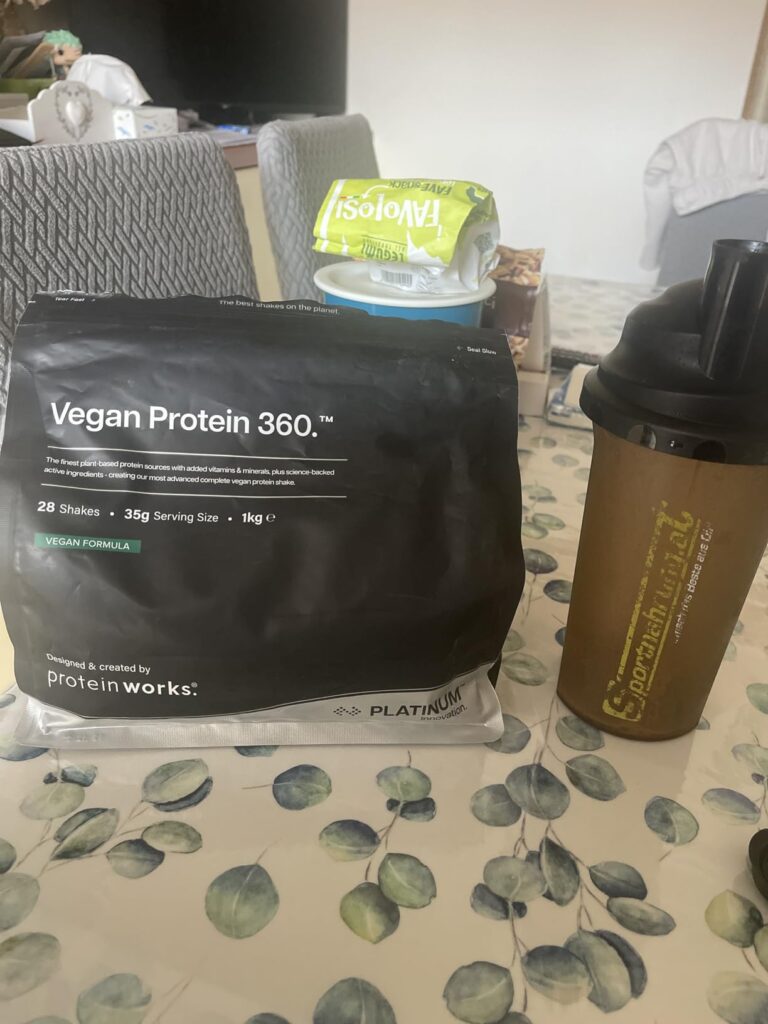 Un sachet de Vegan Protein 360 et un shaker, avec un emballage de fromage frais en arrière-plan.