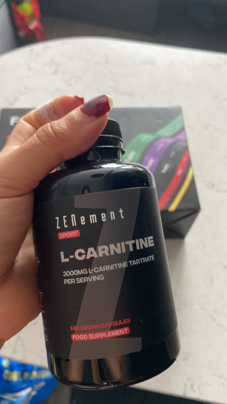 Une main tient le flacon de L-Carnitine, le montrant de face avec l'étiquette principale. Un emballage coloré est visible en...