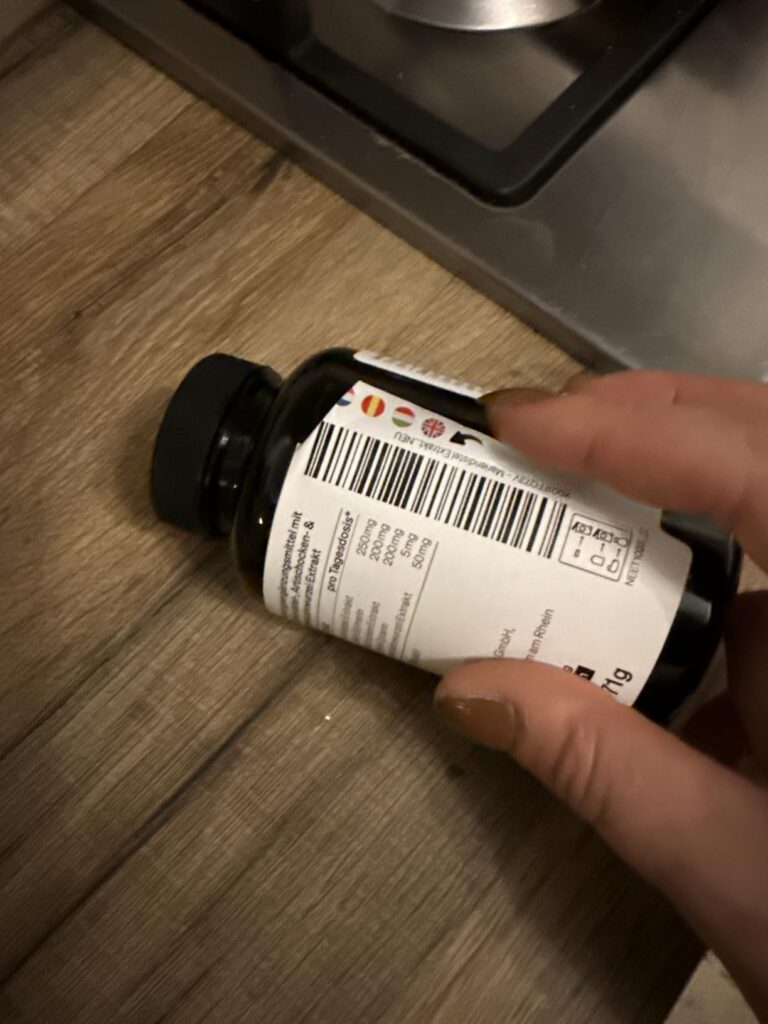 Flacon du produit posé sur le côté, à la main. L'arrière du flacon est visible, avec un code-barres et des icônes de pays.
