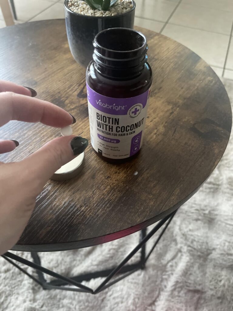 Flacon de biotine Vitabright ouvert sur une table basse, avec un comprimé sorti. Même flacon que le visuel 0 et 1.