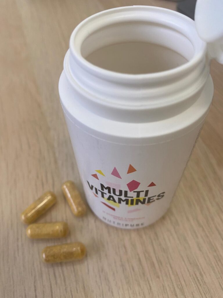 Bouteille de multivitamines ouverte avec des gélules à côté sur une surface en bois clair.