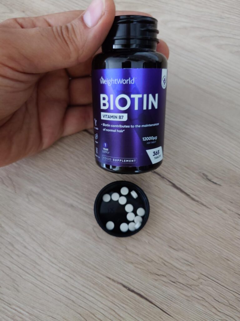 Une main tient le flacon de Biotine, avec le couvercle posé à côté et quelques comprimés blancs visibles.
