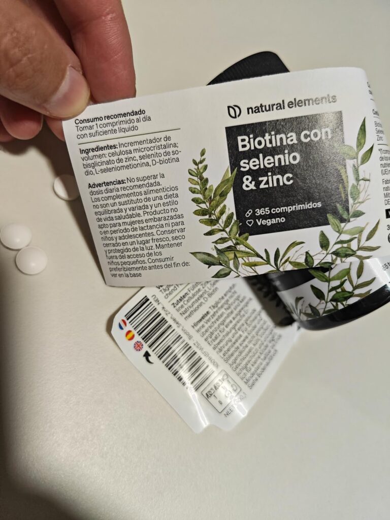 L'image montre le flacon de biotine avec son étiquette décollée, révélant des informations en dessous. Deux comprimés sont po...