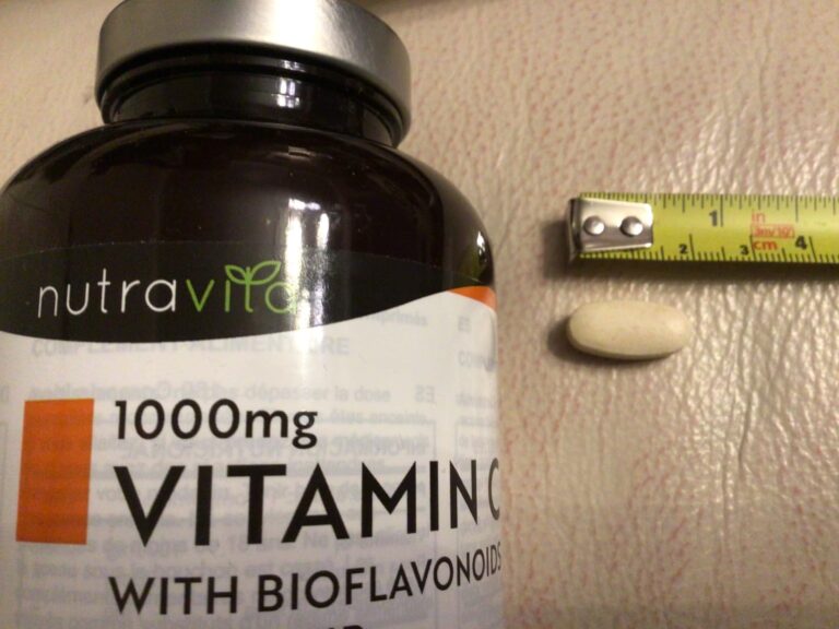 Une gélule de vitamine C à côté d'un ruban à mesurer, avec le flacon en arrière-plan. Le ruban à mesurer est de 15 mm.