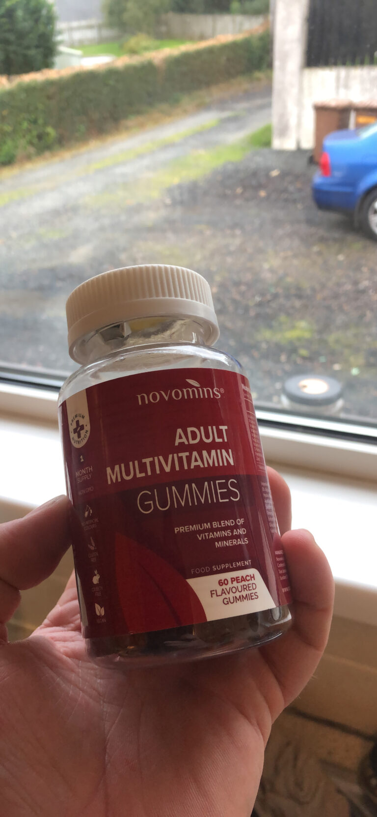 Une main tenant le flacon de "Adult Multivitamin Gummies" de Novomins devant une fenêtre, avec un aperçu de l'extérieur.