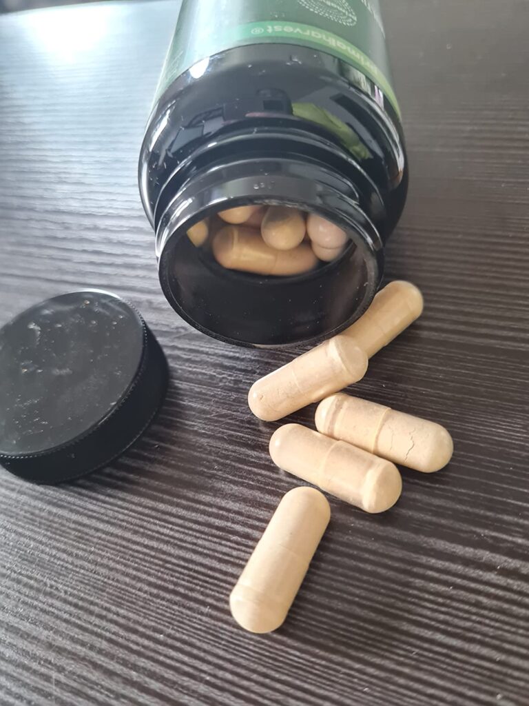 Bouchon de la bouteille ouvert, avec des gélules qui s'échappent, sur une table sombre. La bouteille est la même que dans l'i...