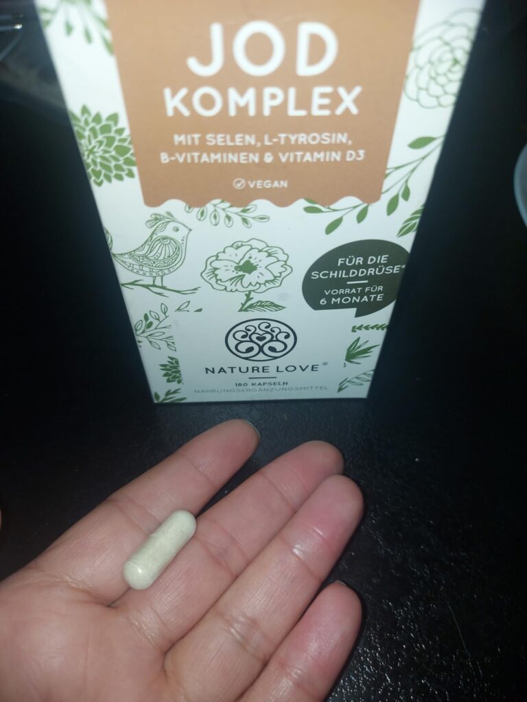 Une capsule de Jod Komplex dans une main, avec l'emballage en arrière-plan.