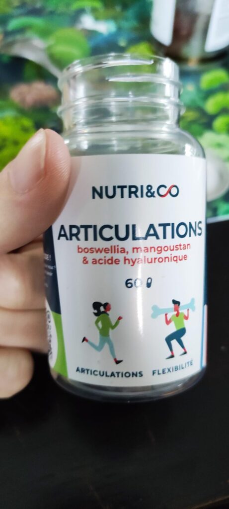 Flacon de compléments alimentaires Nutri&Co, formule articulations, tenu dans une main sur un fond flou qui ressemble à du fe...