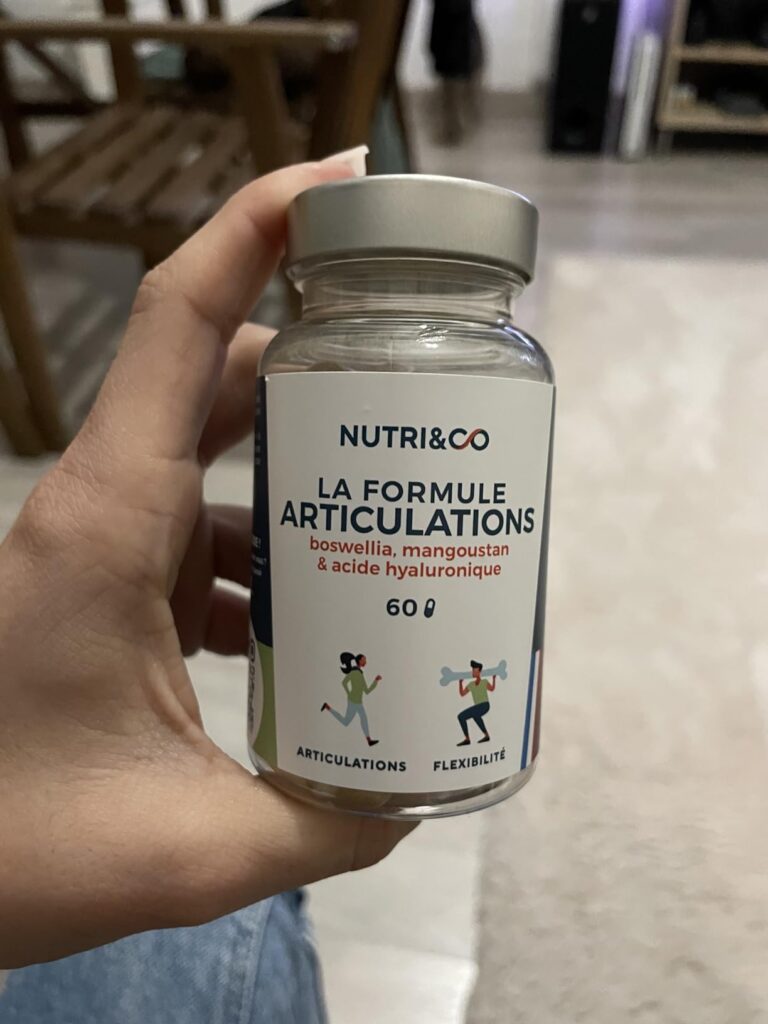 Flacon de compléments alimentaires Nutri&Co, formule articulations, tenu dans une main sur fond flou d'intérieur