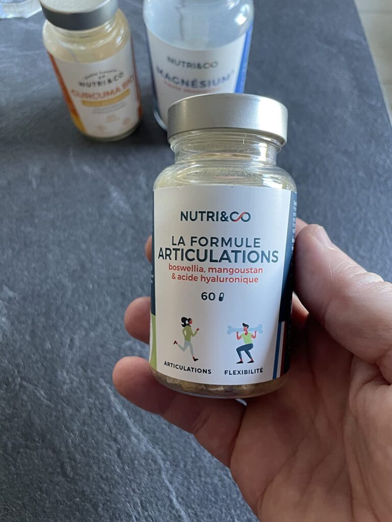Flacon de compléments alimentaires Nutri&Co, formule articulations, tenu dans une main sur un plan de travail, avec deux autr...