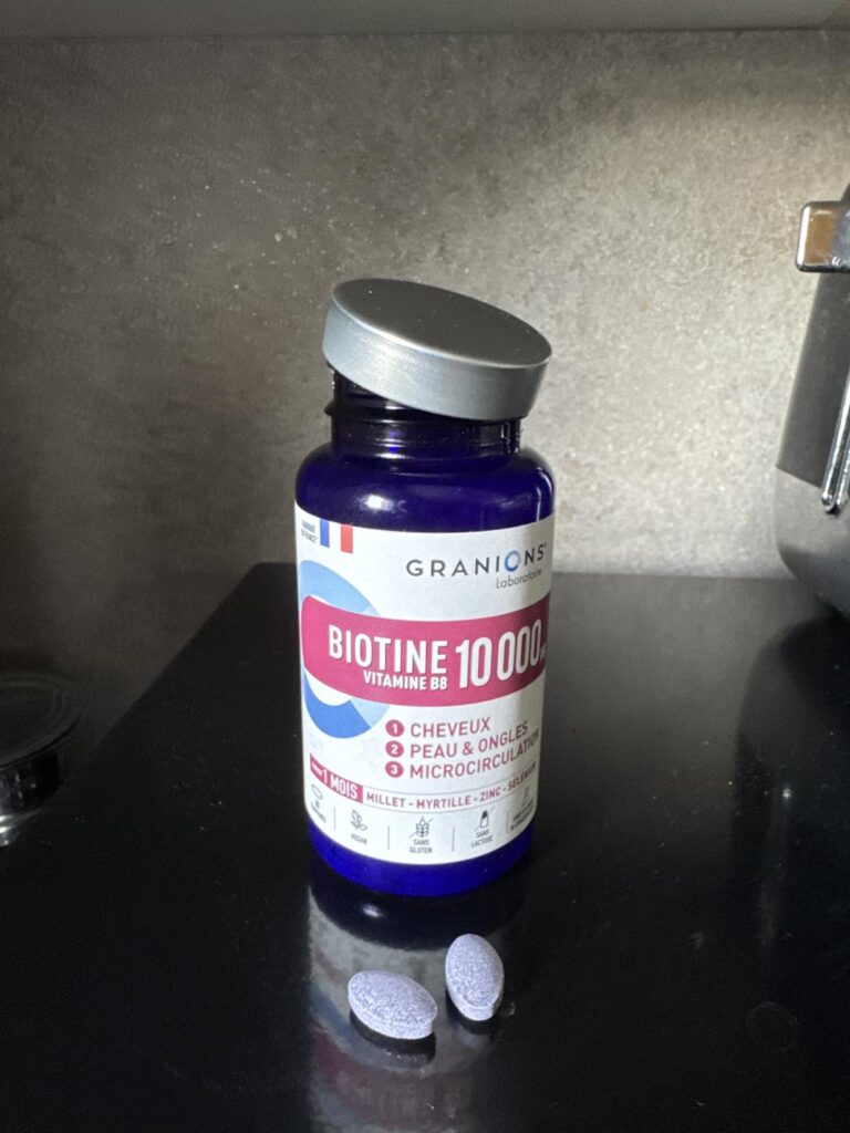 Flacon bleu de Biotine 10 000 mcg sur une surface noire, avec deux comprimés à côté.