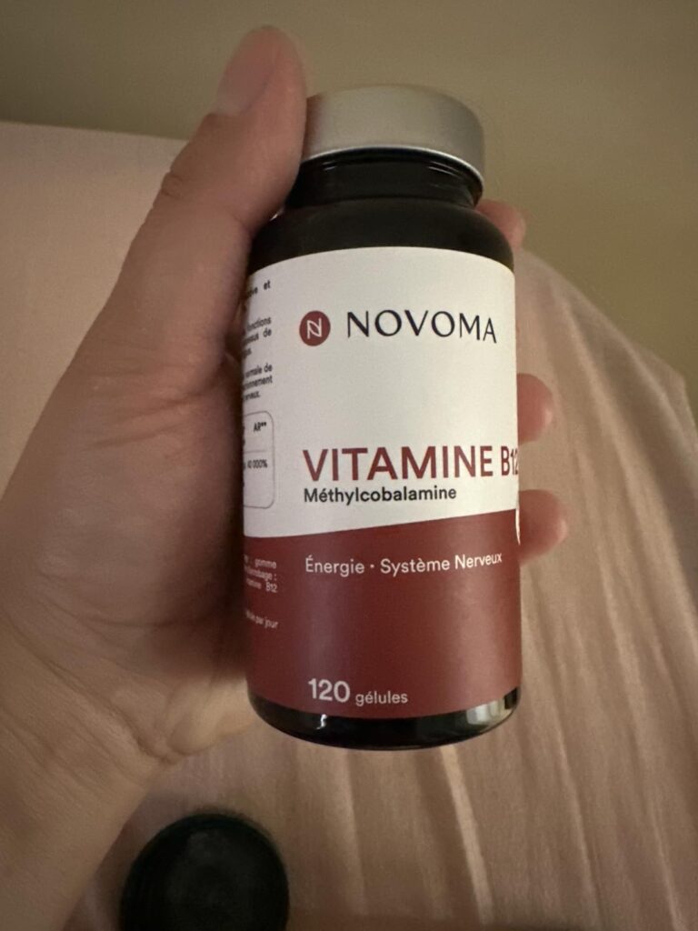 Flacon de compléments alimentaires, tenu à la main, sur fond clair. On peut lire "Novoma Vitamine B12".