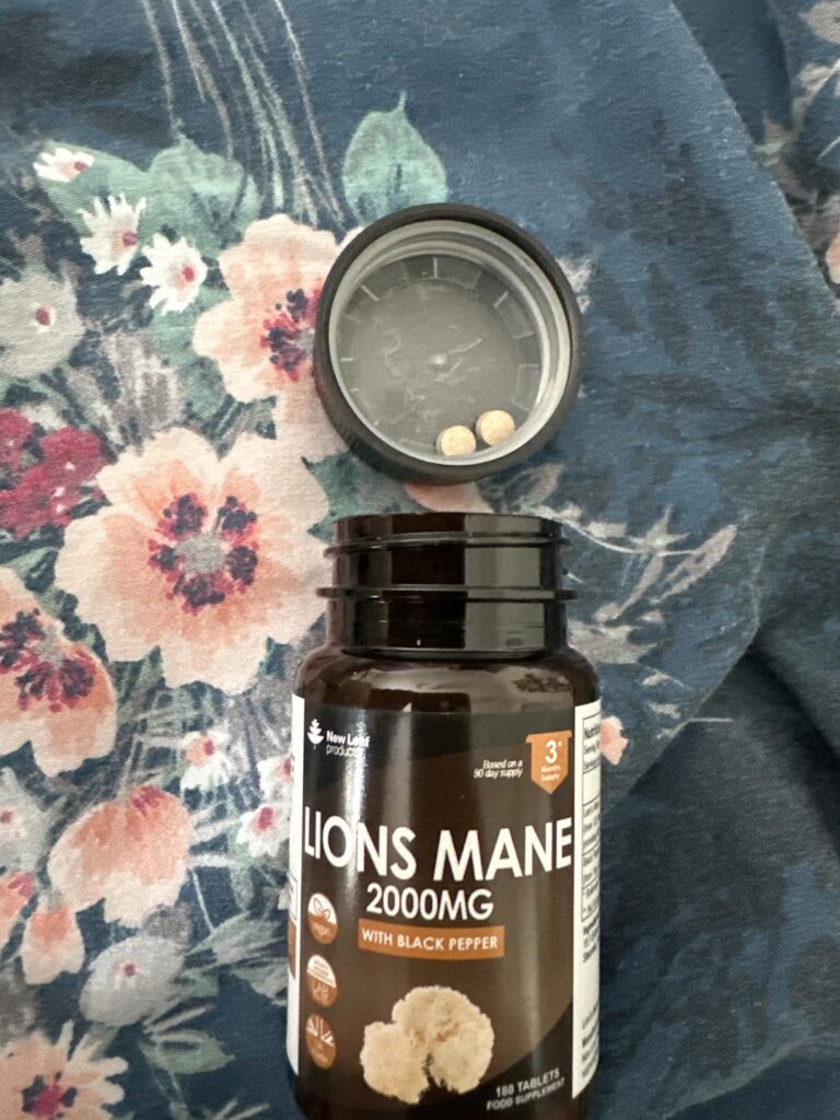 Le flacon de compléments alimentaires ouvert, avec le bouchon à côté, posé sur un tissu à motifs floraux. Deux gélules sont v...