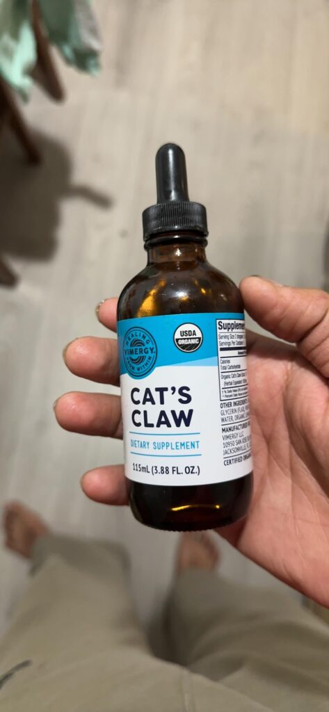 Bouteille de Cat's Claw tenue à la main, avec un compte-gouttes. La bouteille est sur un fond de tissu clair, probablement un...