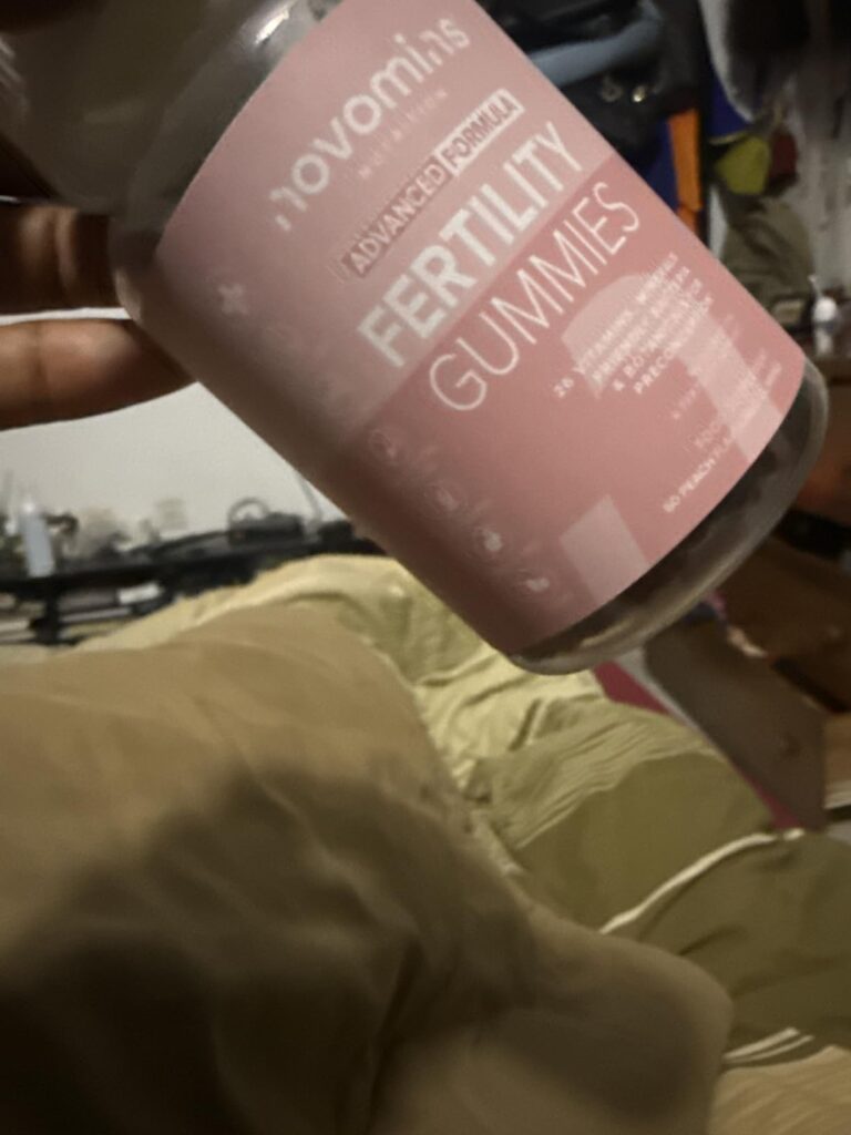 Bouteille de Novomins Fertility Gummies tenue à la main, sur fond flou. Texte clair sur la bouteille : Novomins, Advanced For...