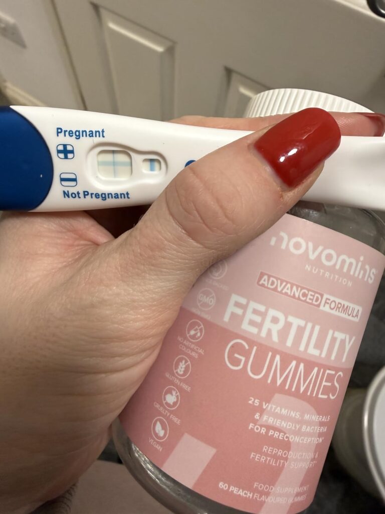 Main tenant une bouteille de Novomins Fertility Gummies et un test de grossesse avec deux lignes bleues et l'indication 'Preg...