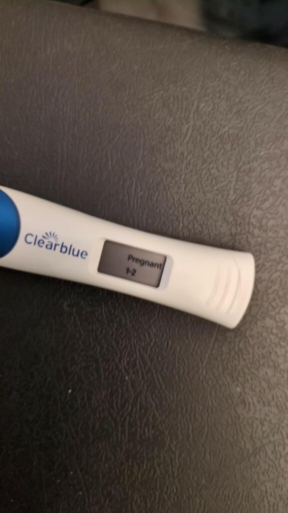 Test de grossesse Clearblue sur fond sombre. L'écran affiche 'Pregnant 1-2'.