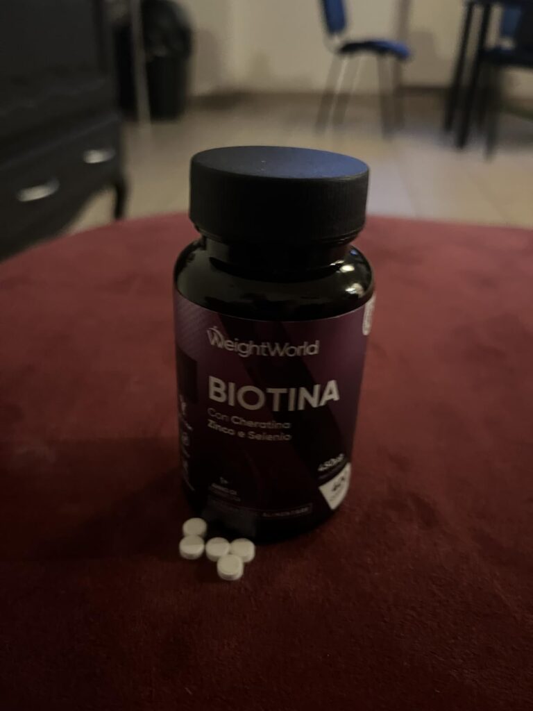 Le flacon de "Biotina" avec quelques comprimés blancs étalés sur une surface rouge. La photo est légèrement floue en arrière-...