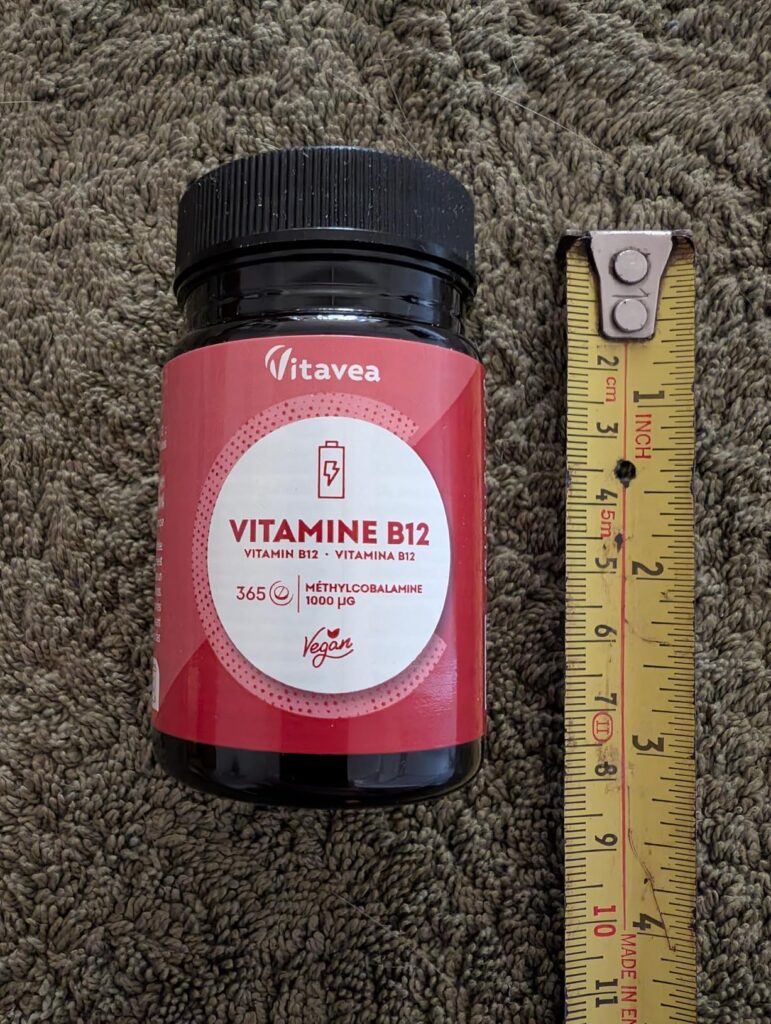 Flacon de vitamine B12 posé à côté d'un mètre-ruban, vue de face. Semblable au visuel 0 mais avec un design d'étiquette légèr...
