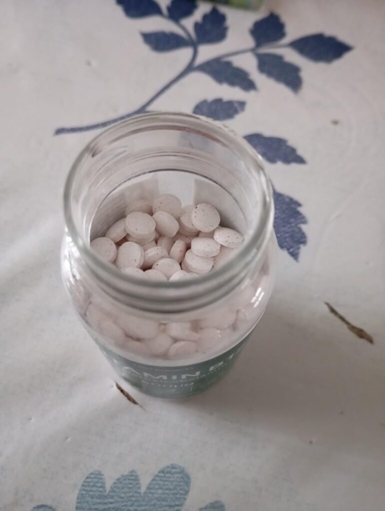 Vue de dessus d'un pot en verre rempli de comprimés ronds et blancs, posé sur une nappe blanche et bleue.