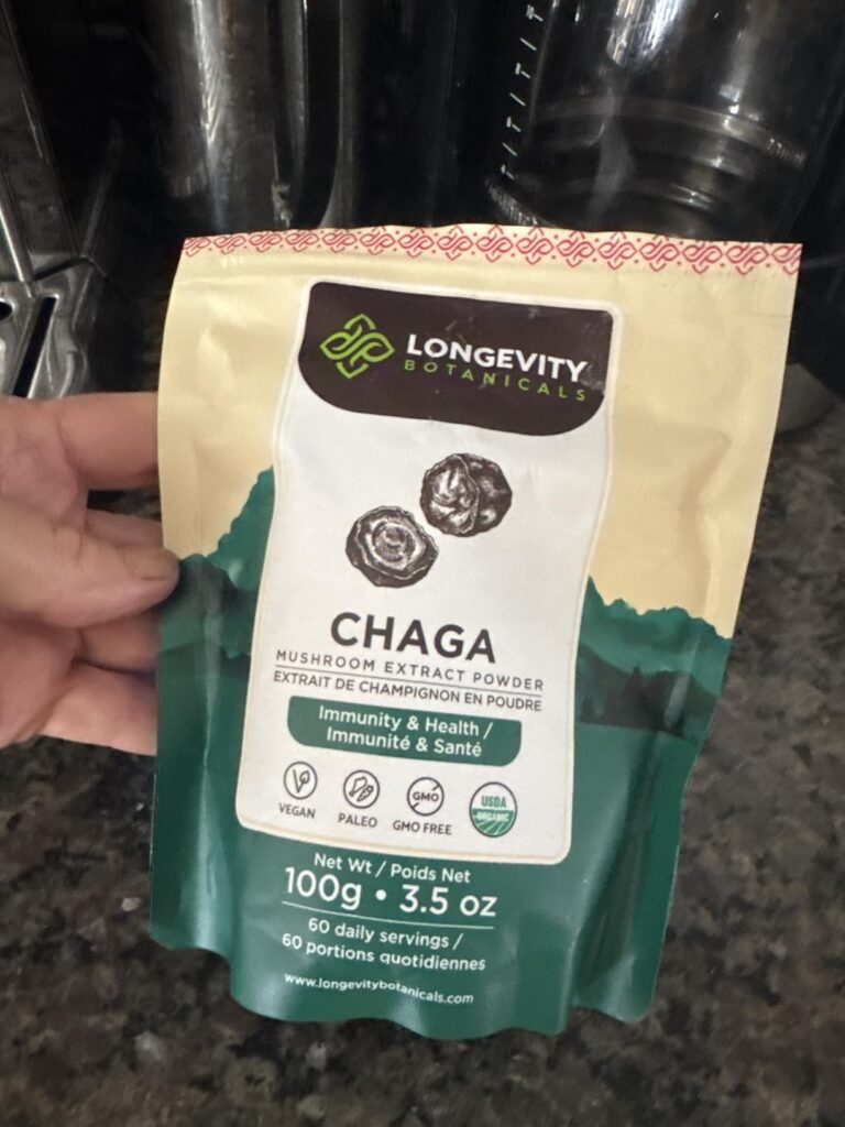 Sachet de la marque Longevity Botanicals, produit Chaga, tenu dans une main. L'arrière-plan est flou, on distingue des objets...