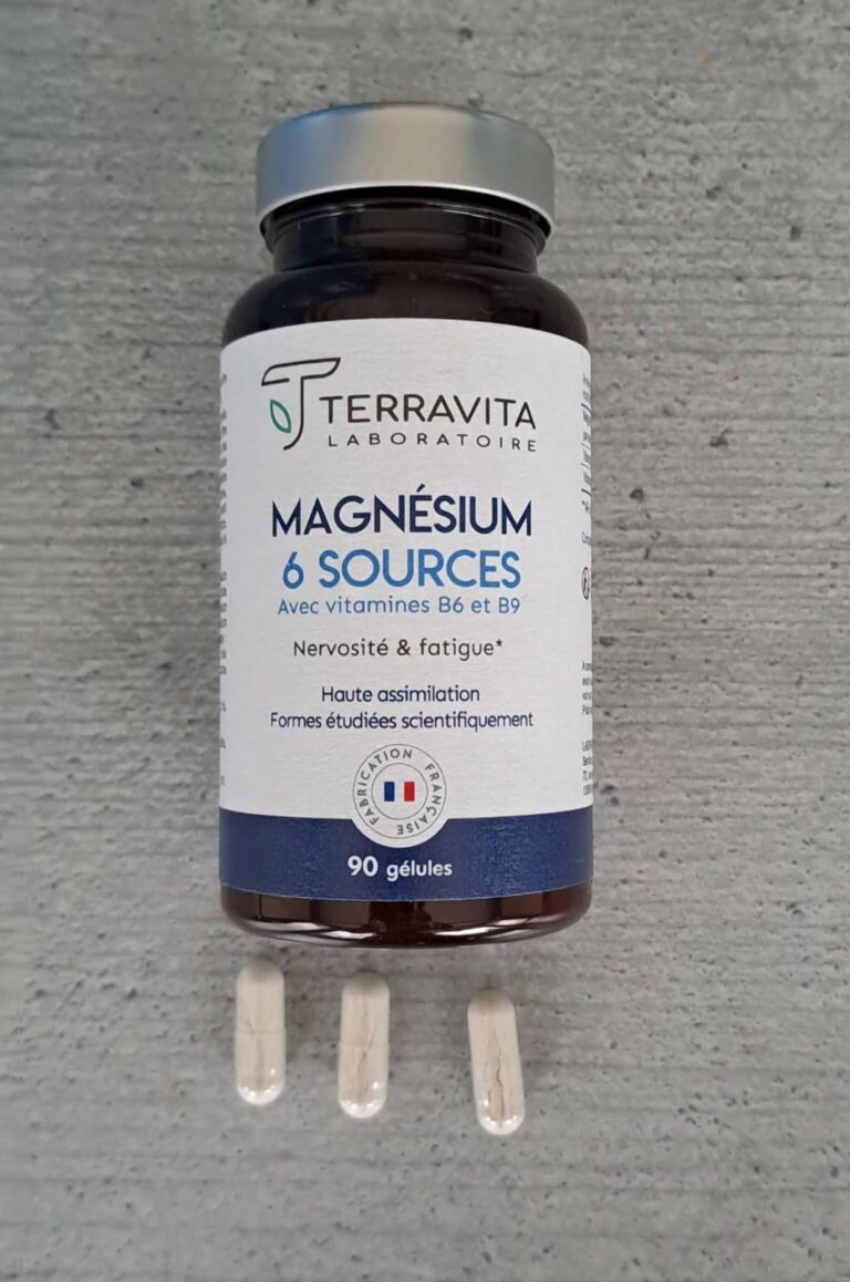 Le flacon du complément alimentaire au magnésium en gros plan, posé sur une surface grise. Trois gélules sont devant le flaco...