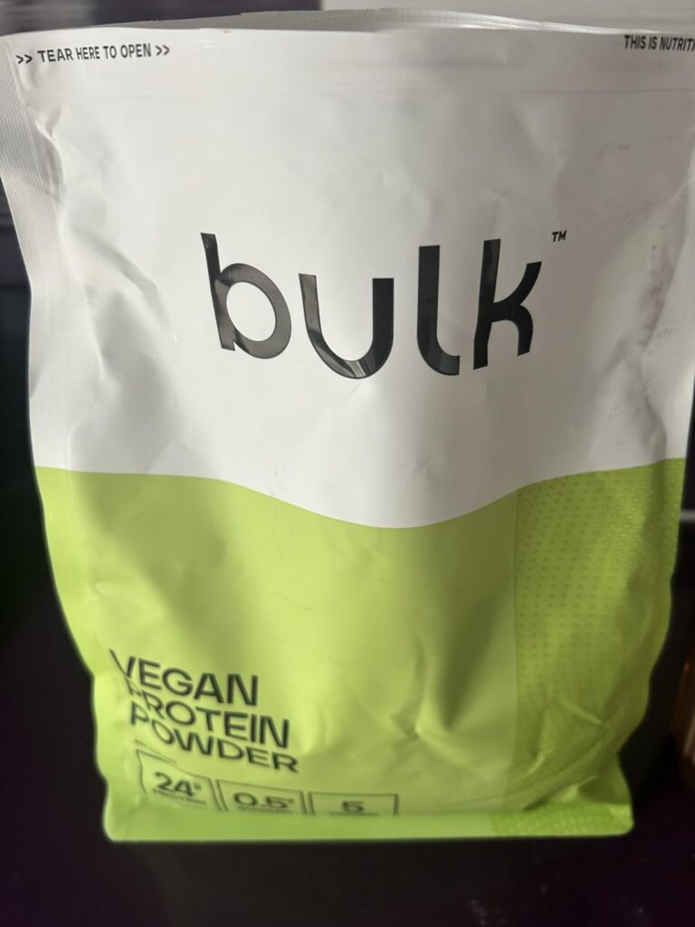 Vue de face du sachet de poudre protéinée Bulk, posé sur une surface sombre. On voit le logo Bulk, la mention Vegan Protein P...