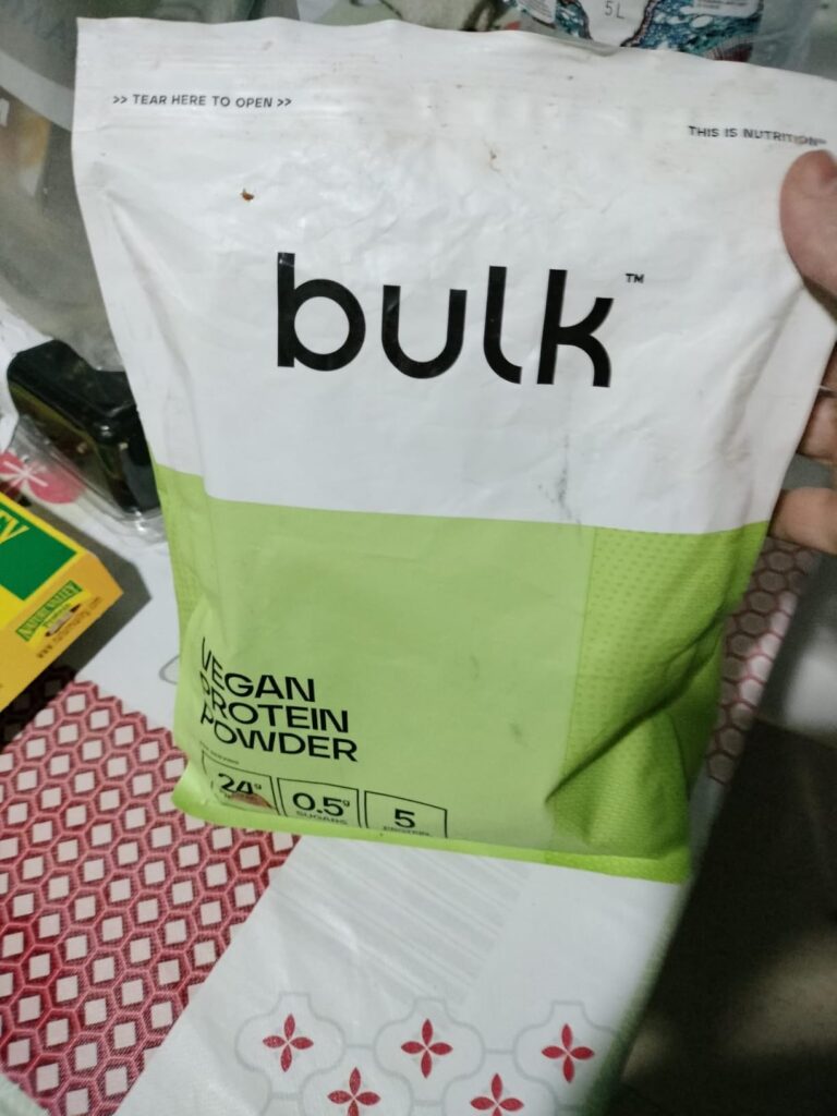 Vue de face du sachet de poudre protéinée Bulk, incliné sur une table avec un fond en tissu rouge et blanc. On voit le logo B...