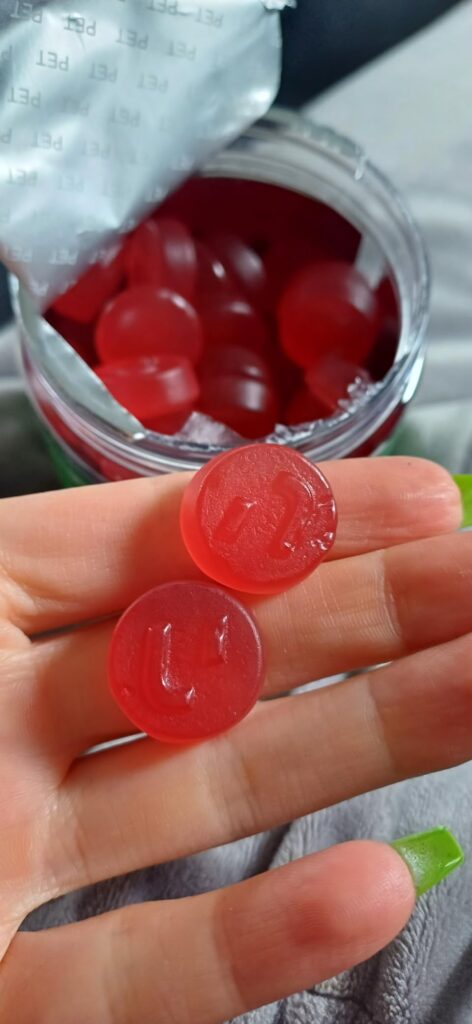 Deux gummies rouges tenus dans une main, avec le pot ouvert en arrière-plan.