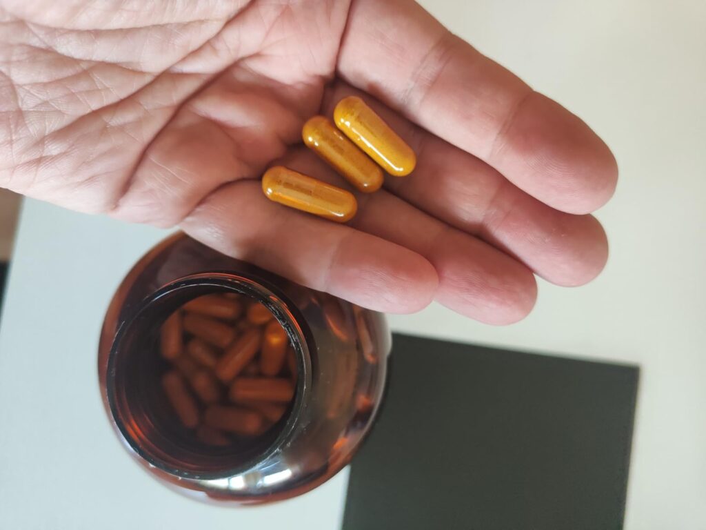 Main tenant quatre capsules jaunes, au-dessus de la bouteille ouverte et pleine de capsules. Les capsules sont jaunes.