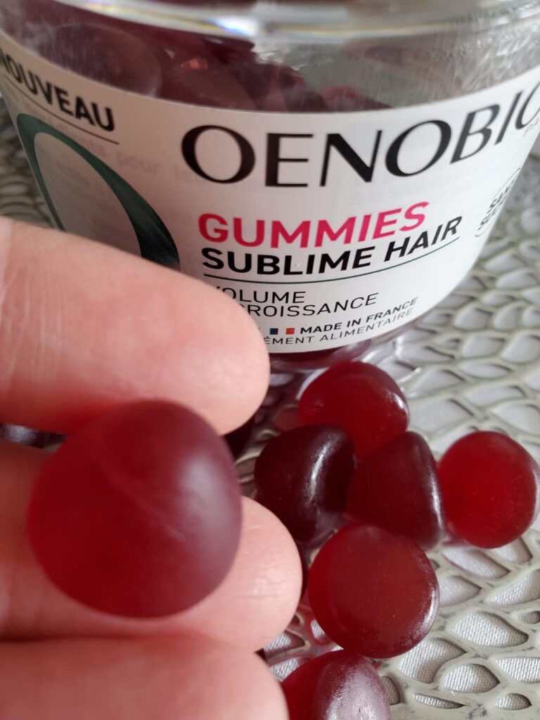 Un bocal de "Gummies Sublimes Hair" de la marque Oenobiol, avec une pastille en résine à la main et d'autres dans le bocal.