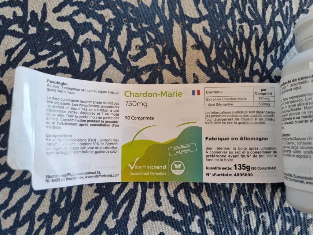 L'étiquette du produit sur fond de tissu imprimé, avec un flacon blanc partiellement visible sur la droite. L'étiquette est e...