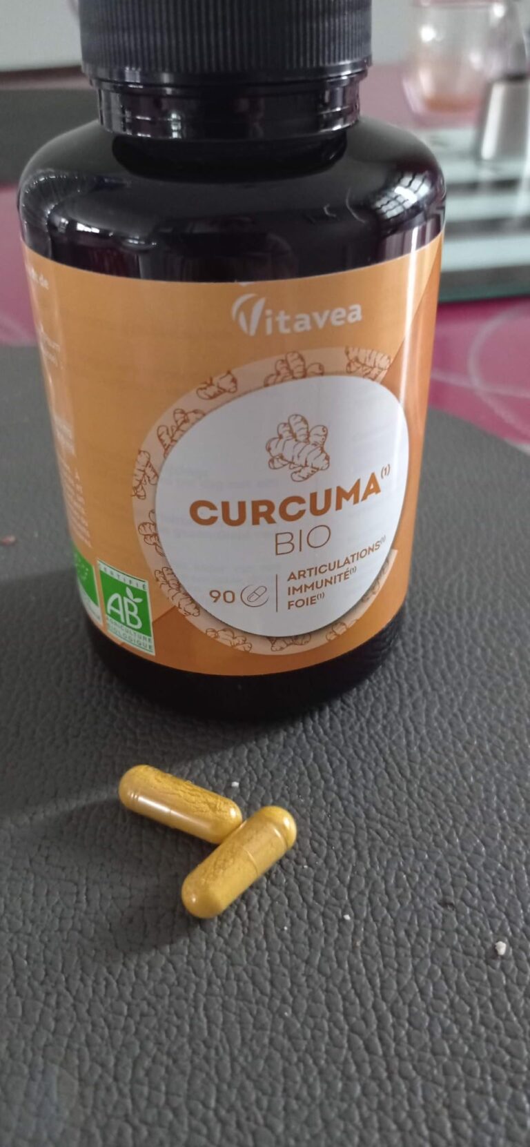 Un flacon de curcuma en arrière-plan, avec deux gélules jaunes au premier plan sur une surface grise. Le flacon porte la marq...