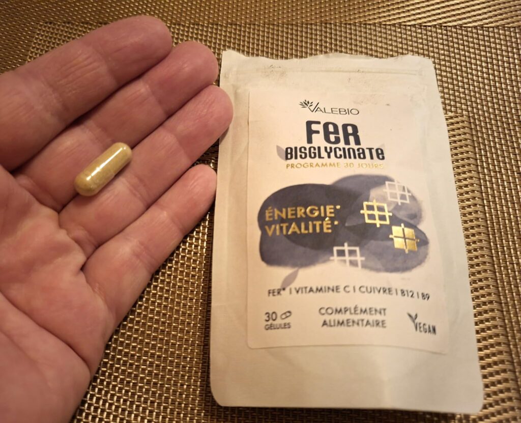 Une gélule tenue dans la paume d'une main, et le sachet sur une surface tressée foncée. Angle différent du visuel 1.