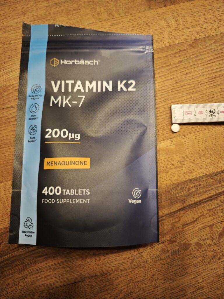 Sachet de vitamine K2 Horbäach et un comprimé posé à côté d'une règle.