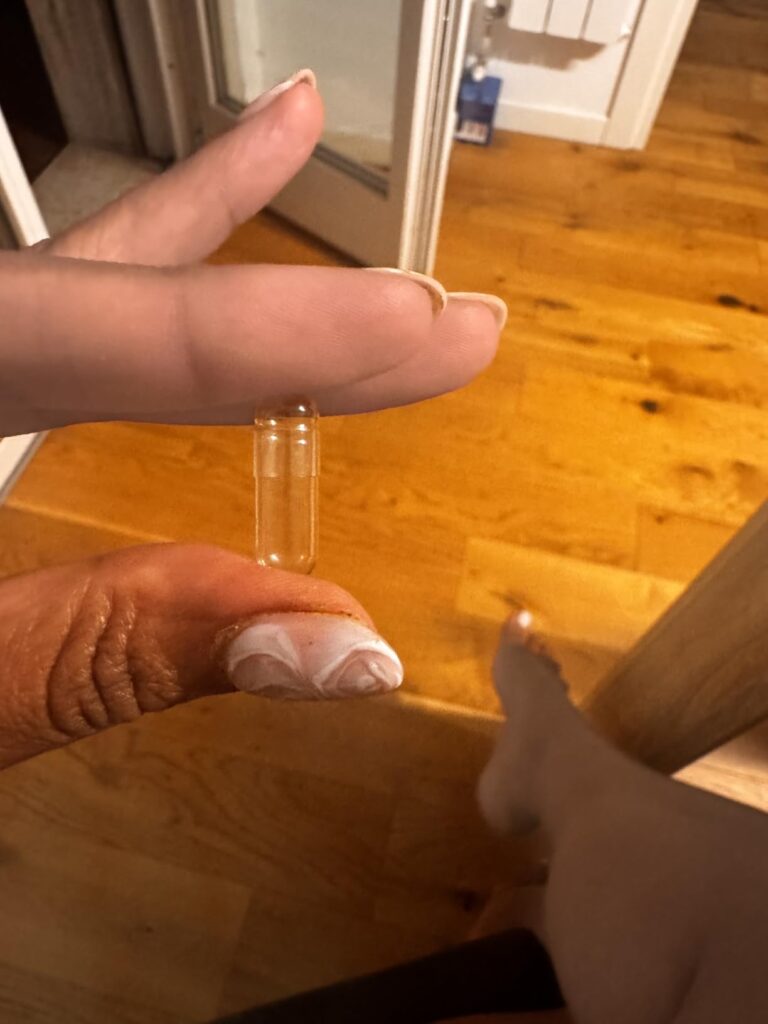 Une capsule vide tenue entre deux doigts, avec un arrière-plan en bois et un pied visible en bas à droite.